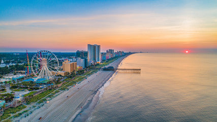 Aerial view of a beach with a ferris wheel at sunset, showcasing coastal skyline and calm ocean waves.